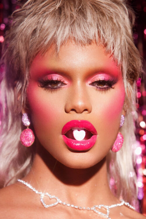 Close-up of a young woman with pale blonde hair and pink makeup holding a candy heart in her mouth, against a shiny pink background.