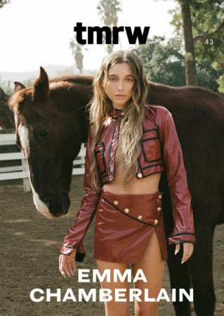 Emma Chamberlain wearing a red patent leather cropped jacket and miniskirt standing next to a dark brown horse outdoors.