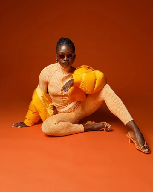 An adult woman poses in matching knit top, leggings, high heels, and puffy jacket, all orange, set against an orange backdrop.