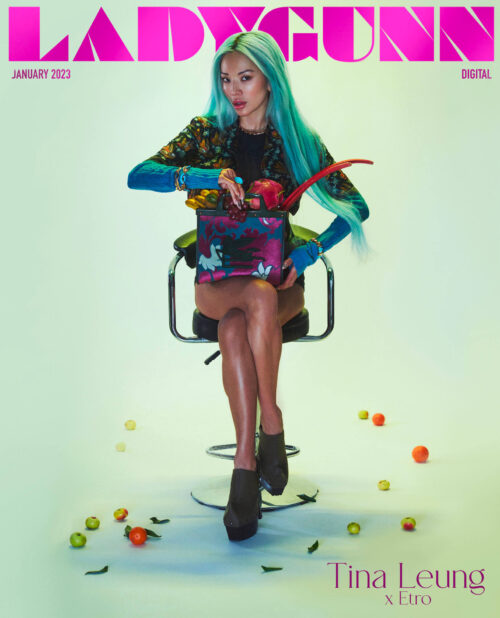 Magazine cover featuring a model with aqua hair sitting on a chair, holding a container with flowers and fruit, surrounded by fruit.
