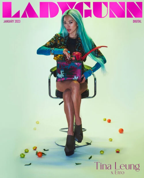 Magazine cover featuring a model with aqua hair sitting on a chair, holding a container with flowers and fruit, surrounded by fruit.