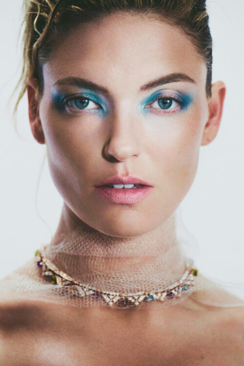 Close-up portrait of a fair-skinned adult female model with blue eyeshadow, a sheer draped collar, and an elaborate jeweled necklace in a white space.