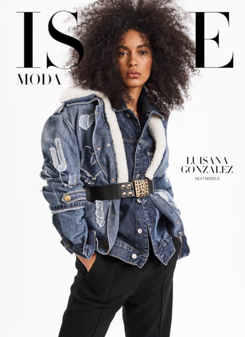Editorial fashion shot of a woman with curly hair wearing a denim jacket and wide belt against a white background in a studio.