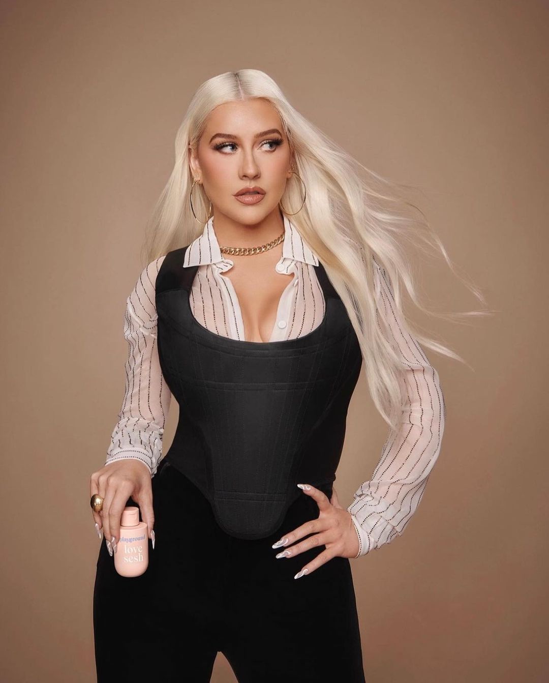 Christina Aguilera poses in a photo studio wearing a black corset, striped shirt, gold jewelry, and holding a skincare product.