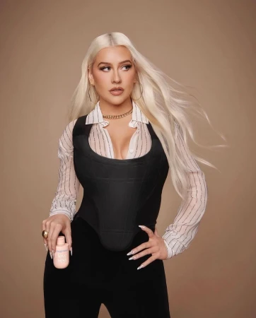 Christina Aguilera poses in a photo studio wearing a black corset, striped shirt, gold jewelry, and holding a skincare product.