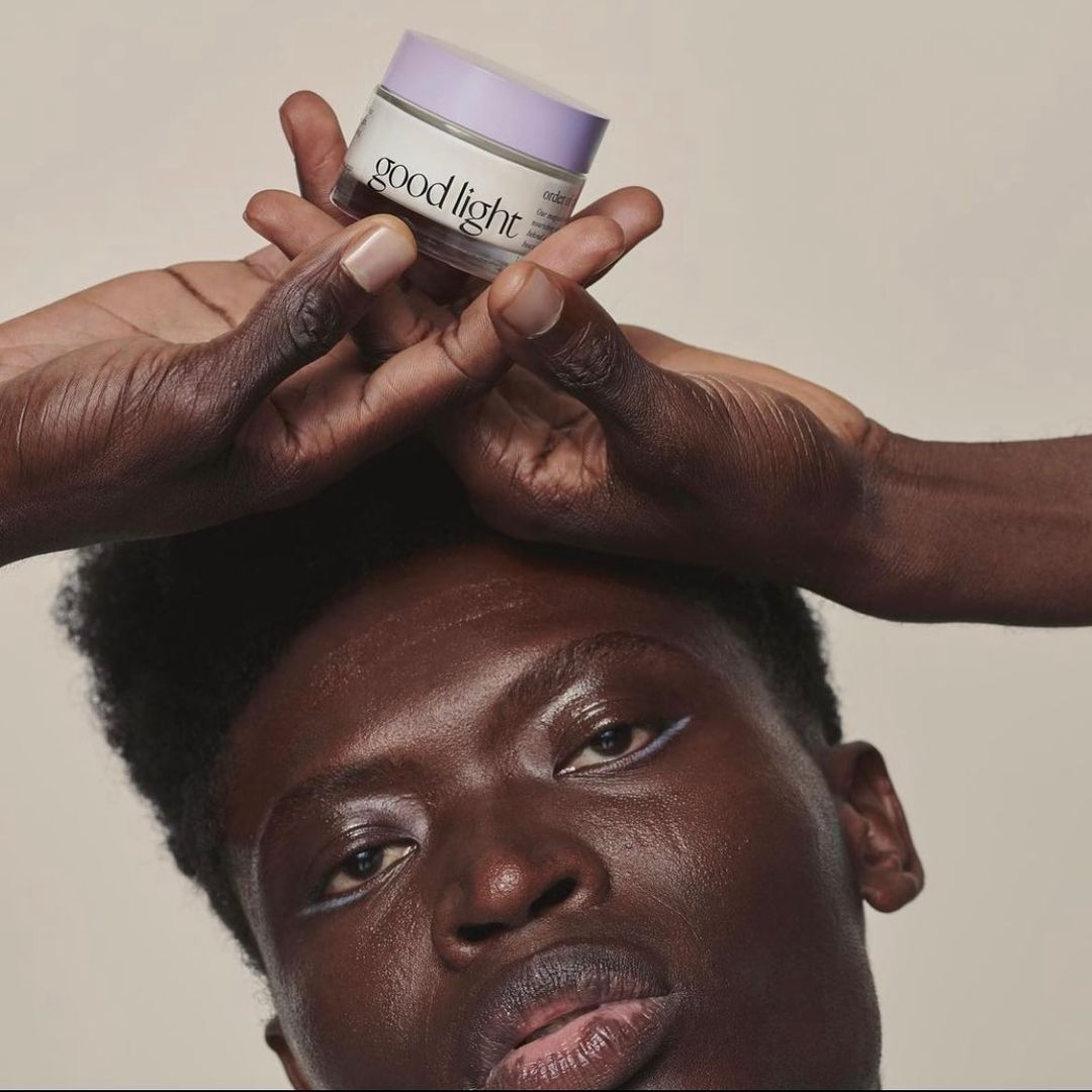 Person holds Good Light skincare jar overhead. African American male with subtle white eyeliner, clean shaven, studio setting.