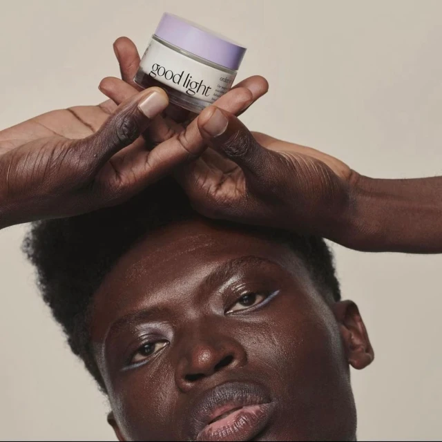 Person holds Good Light skincare jar overhead. African American male with subtle white eyeliner, clean shaven, studio setting.