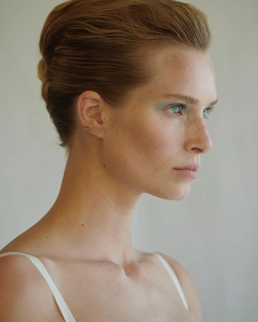 Close-up portrait of a fair-skinned adult woman with short, light brown hair styled back, featuring green makeup around the eyes and fair complexion.