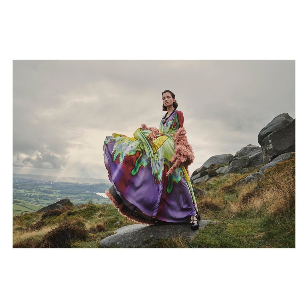 Woman in a colorful dress poses on a rock outcrop in an open landscape with cloudy skies, photographed in medium shot at eye level.