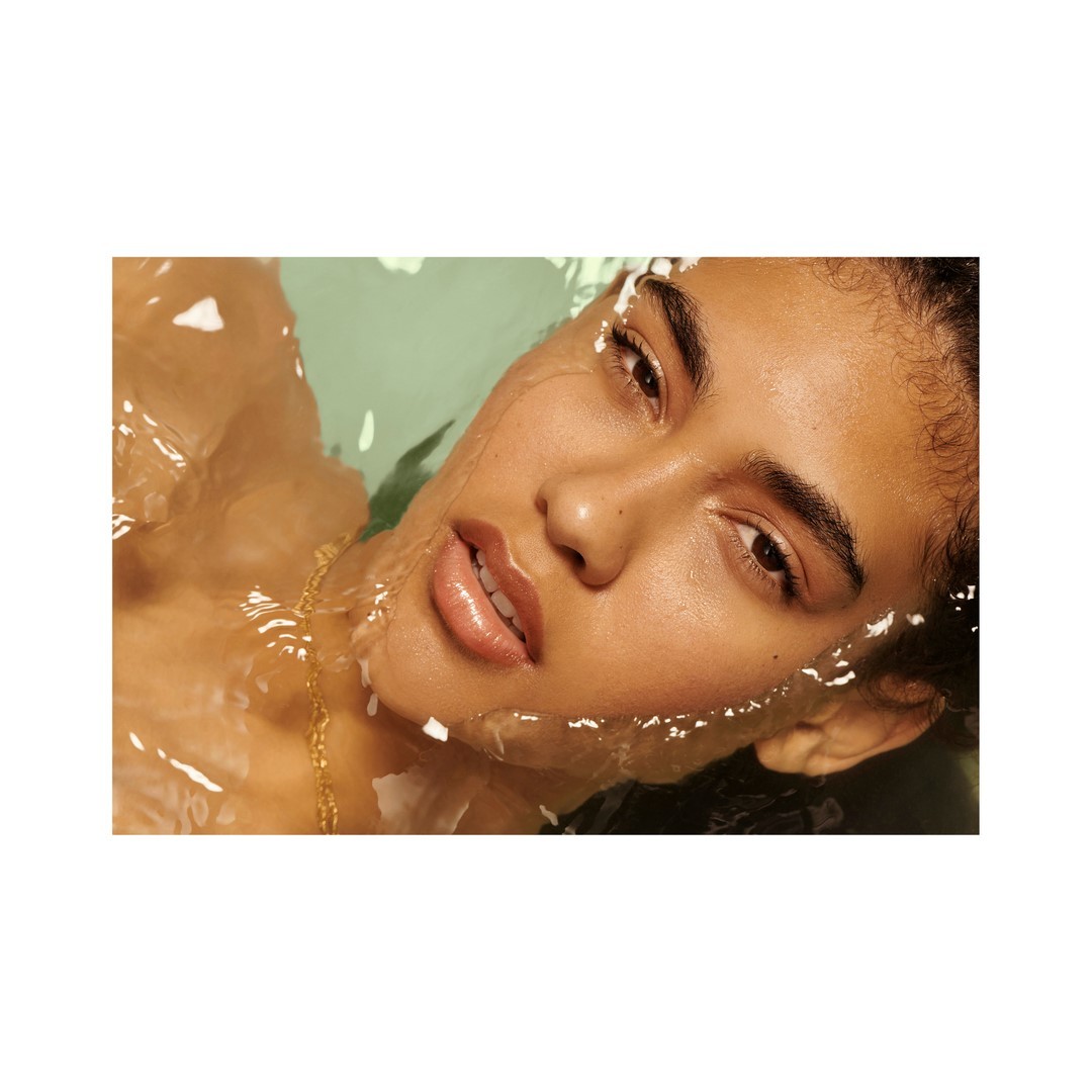 Close-up of a woman with tan skin lying in water, gazing at the camera with open lips, wearing a gold necklace and light makeup.