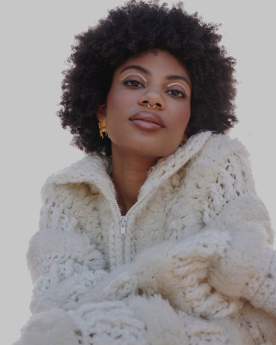 Close-up of a stylish adult woman with Afro hair wearing a white textured jacket and gold earrings against a grey background.