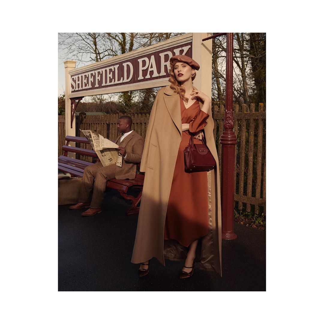 A woman in a dress and trench coat stands at a train station, with a man reading a newspaper on a bench in the background.