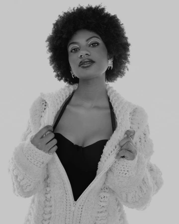 Black and white portrait of a woman with an afro wearing a jacket, standing against a neutral backdrop with modern makeup.