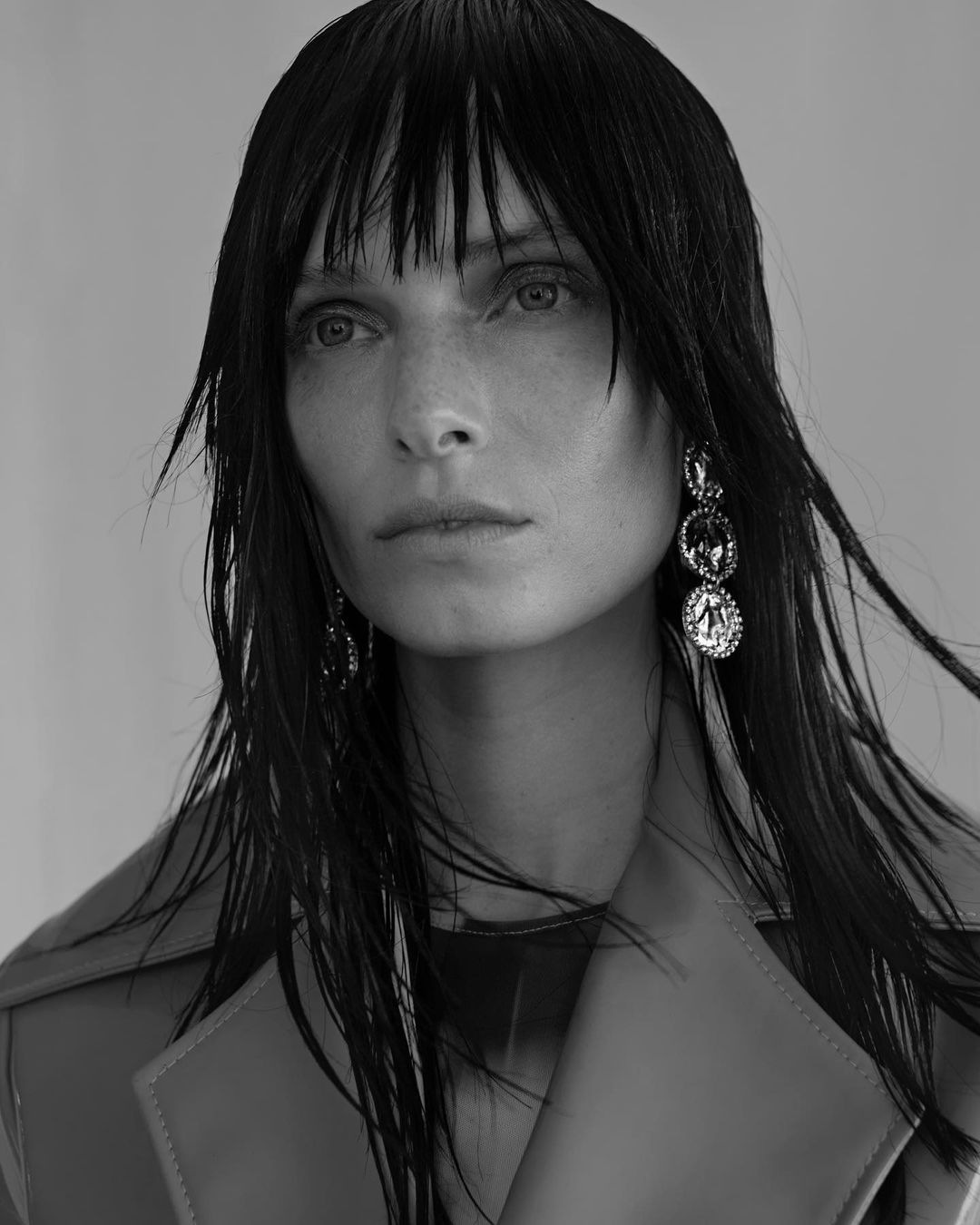 Black and white portrait of a woman with dark, wet hair and wearing a trench coat and jeweled earrings. Fashion and beauty concept.