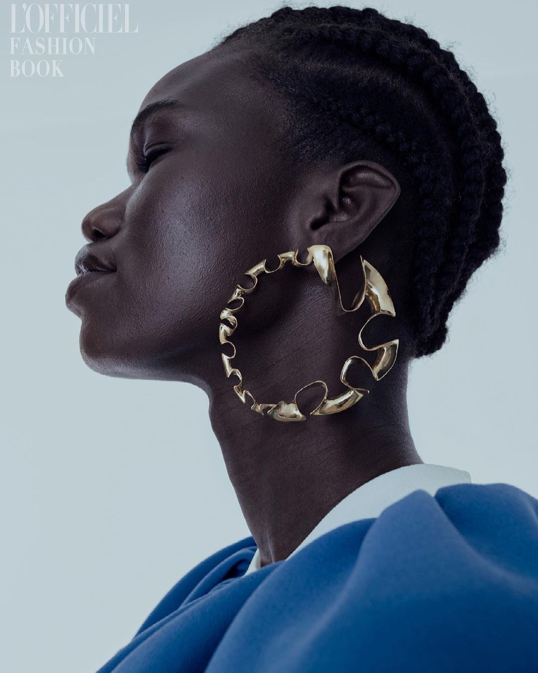 Close-up fashion shot of an African woman with braided hair wearing a gold earring and a blue dress on a light background.