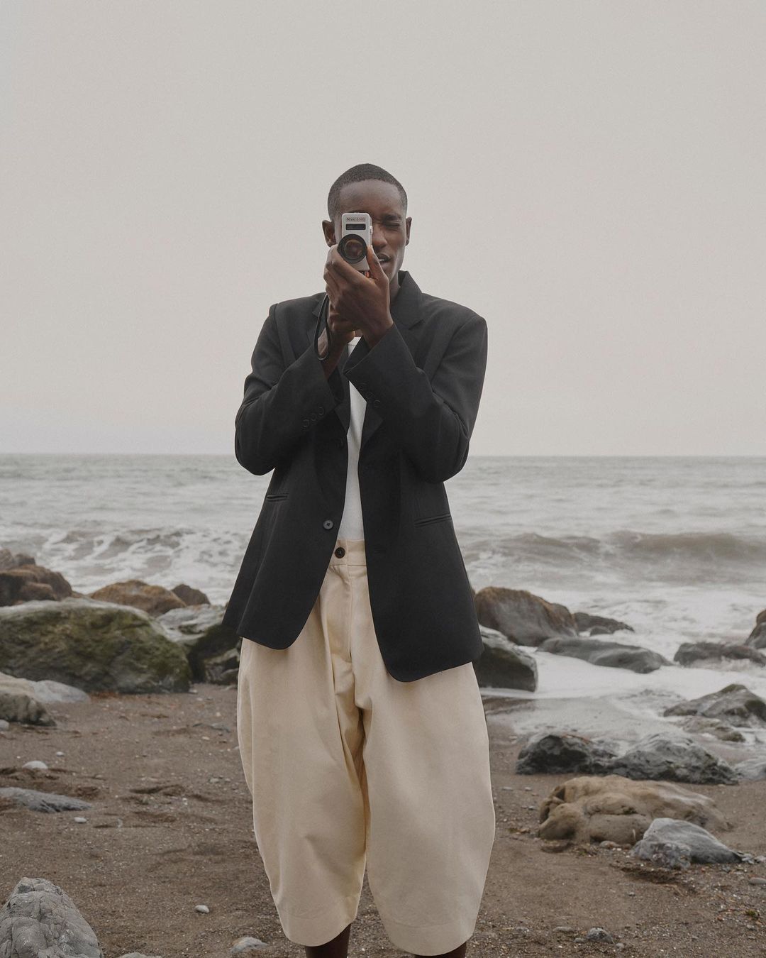 Man films at the beach with vintage camera. Person in dark blazer and light trousers holds camera on cloudy day by the ocean.