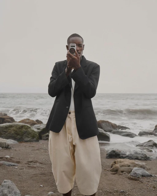Man films at the beach with vintage camera. Person in dark blazer and light trousers holds camera on cloudy day by the ocean.