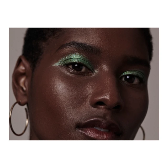 Close-up of a dark-skinned woman with short hair wearing green eyeshadow and gold hoop earrings against a neutral gray backdrop.