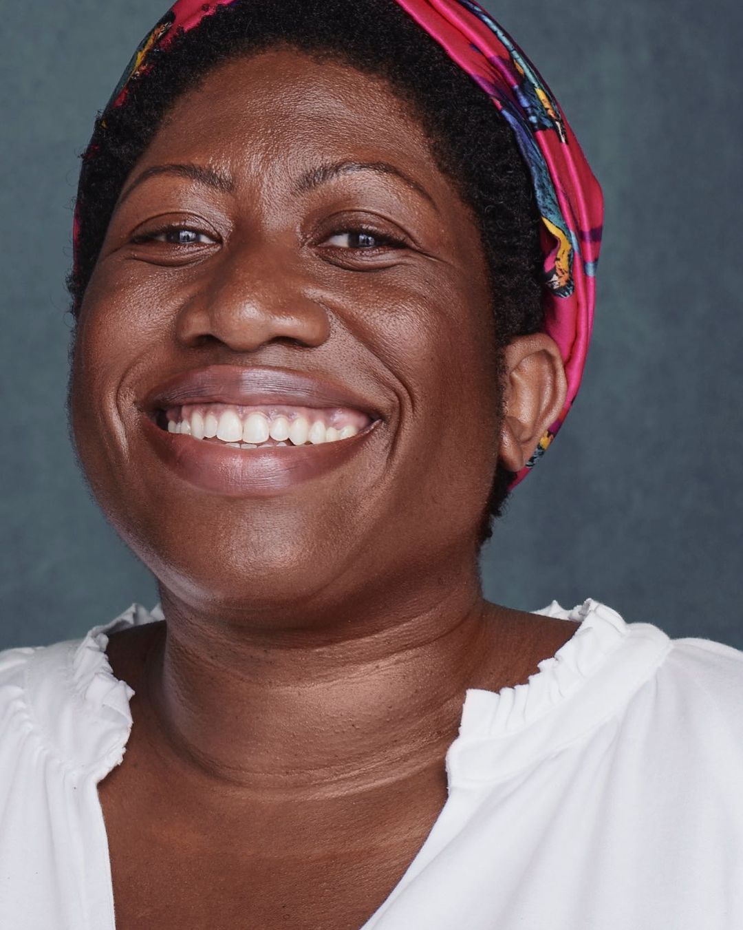 Close-up portrait of a smiling Black woman wearing a colorful headscarf and a white blouse against a neutral blue background.