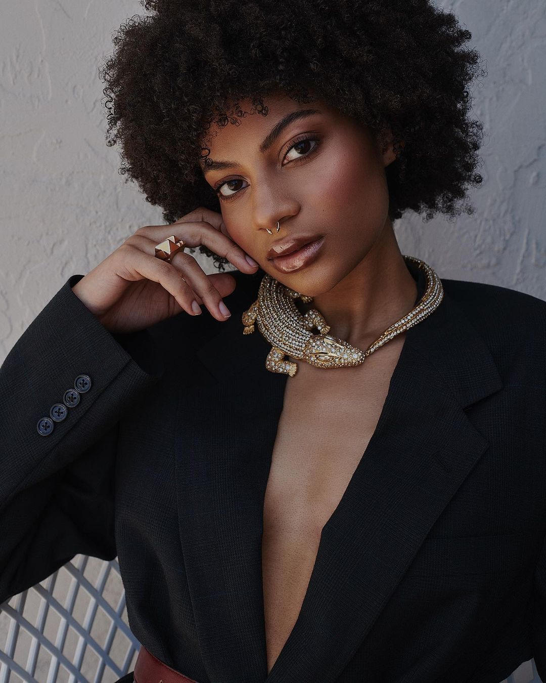 Portrait of a young woman with curly hair wearing a black blazer and an alligator necklace, posing in front of a textured wall.