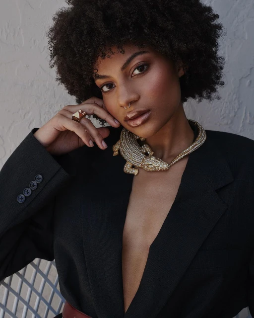 Portrait of a young woman with curly hair wearing a black blazer and an alligator necklace, posing in front of a textured wall.