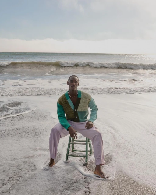 Man in colorful cardigan and light purple pants sits on a chair on a sandy beach with ocean waves and blue sky in the background.