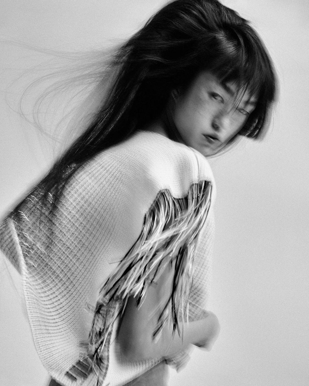 Black-and-white portrait of an Asian woman with dark hair wearing a sweater with fringe, gazing back over her shoulder in a studio setting.