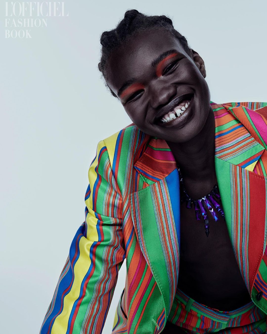Close-up fashion shot of a dark-skinned adult woman smiling, wearing a multicolored striped suit, necklace, and bold red eyeshadow.