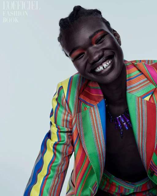 Close-up fashion shot of a dark-skinned adult woman smiling, wearing a multicolored striped suit, necklace, and bold red eyeshadow.