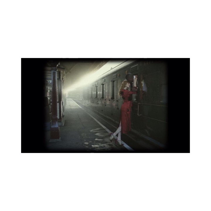 Woman in red dress leans out train window to kiss man on station platform, evocative of 1940s farewell scene with light fog.