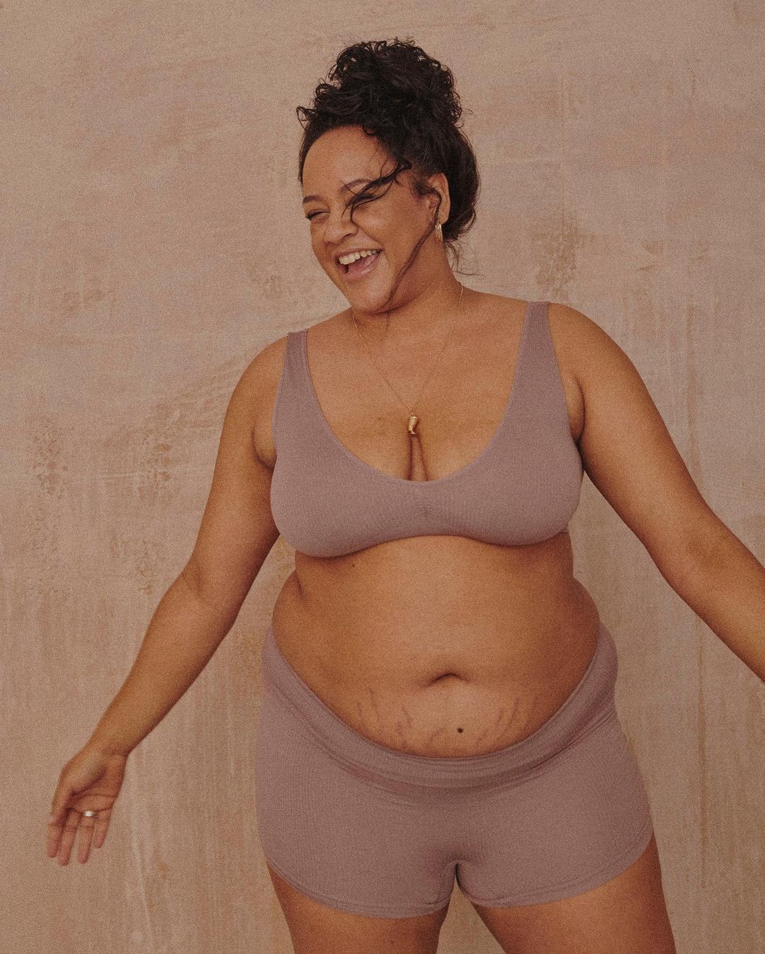 An adult woman smiles in a studio shot, wearing a mauve-colored bra top and shorts, accessorized with a delicate gold necklace.