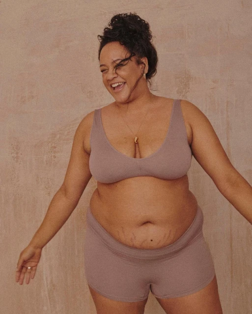 An adult woman smiles in a studio shot, wearing a mauve-colored bra top and shorts, accessorized with a delicate gold necklace. 