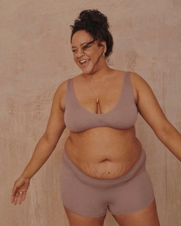 An adult woman smiles in a studio shot, wearing a mauve-colored bra top and shorts, accessorized with a delicate gold necklace. 