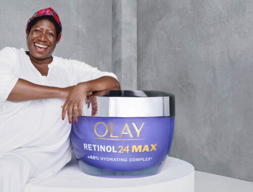 African American woman smiles next to a jar of OLAY Retinol 24 MAX moisturizer in a beauty product advertisement against a gray backdrop.