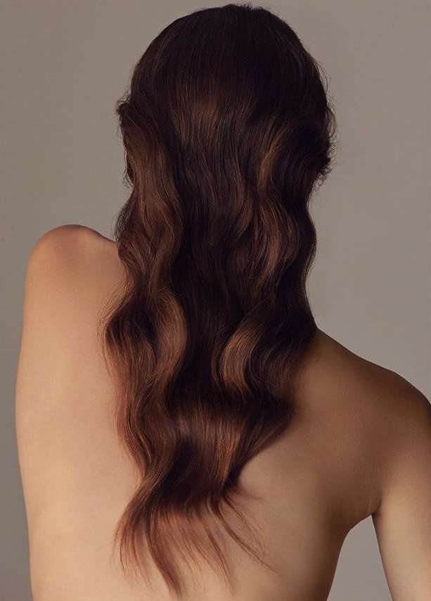 Close-up of an adult female with long, wavy auburn hair cascading down her bare back, set against a neutral beige backdrop.