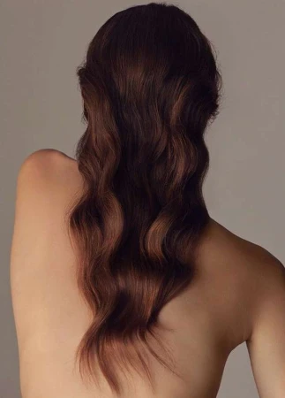 Close-up of an adult female with long, wavy auburn hair cascading down her bare back, set against a neutral beige backdrop.