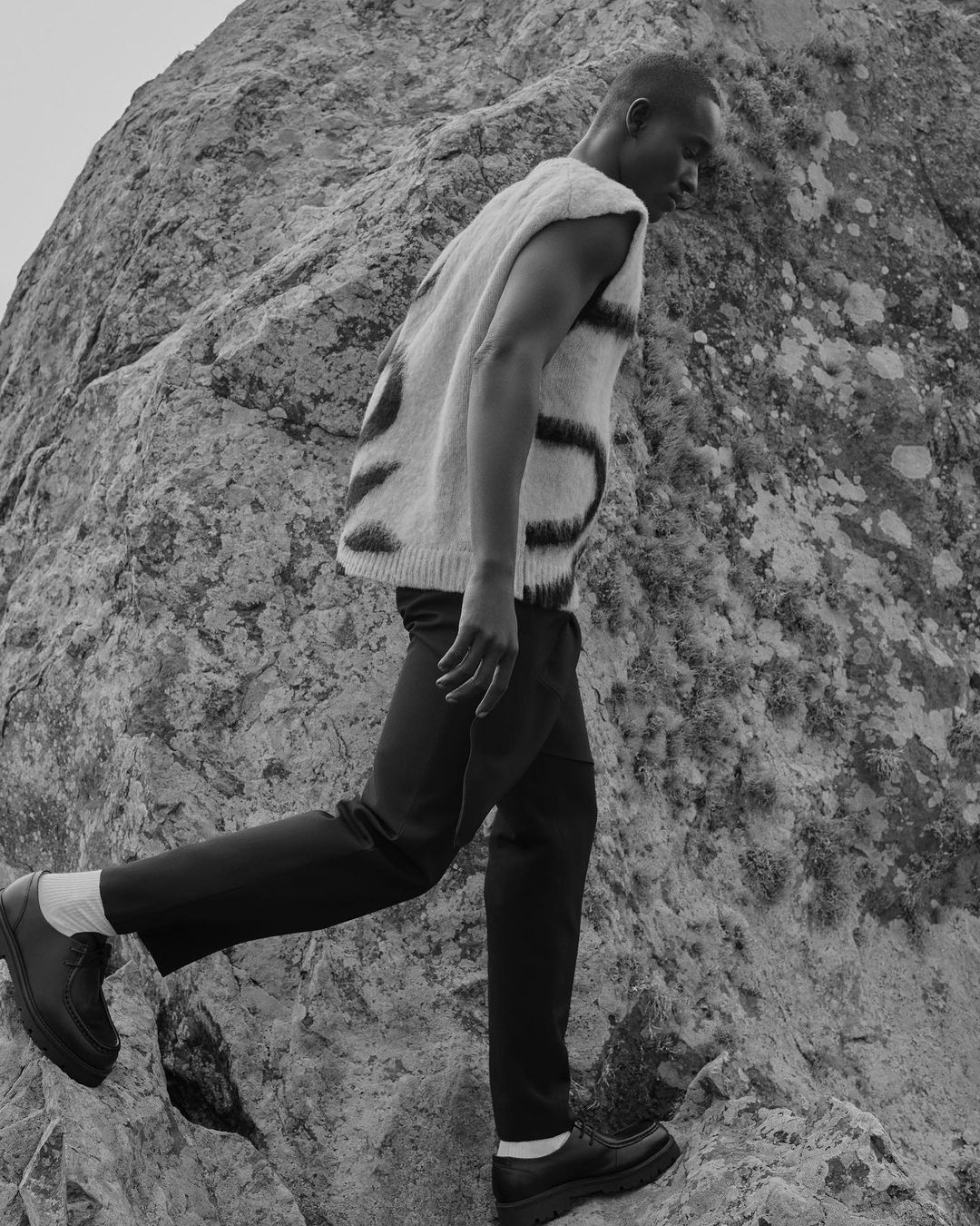 Black man climbing a rocky terrain wearing a patterned vest, dark trousers, and chunky leather shoes.