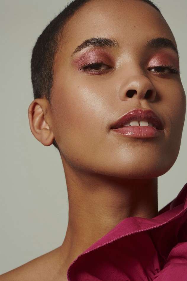 Close-up portrait of an adult female with short hair and pink eyeshadow, wearing a fuchsia-colored garment, against a neutral background.