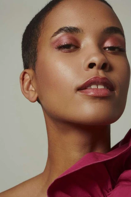 Close-up portrait of an adult female with short hair and pink eyeshadow, wearing a fuchsia-colored garment, against a neutral background.