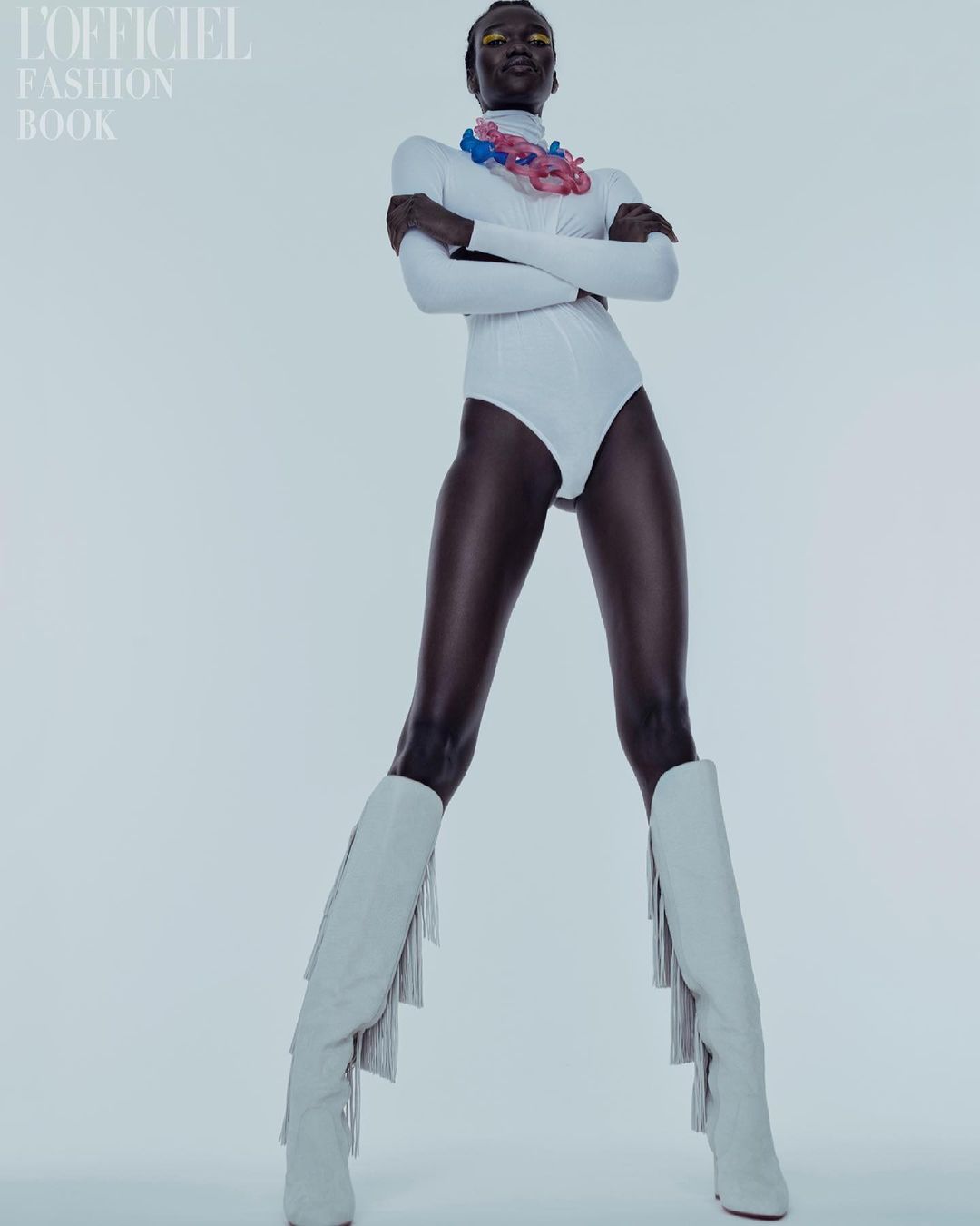 Full-length studio shot of a dark-skinned woman in a white bodysuit and heeled boots, posing confidently against a plain background.