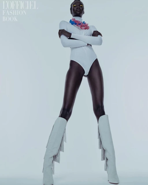 Full-length studio shot of a dark-skinned woman in a white bodysuit and heeled boots, posing confidently against a plain background.