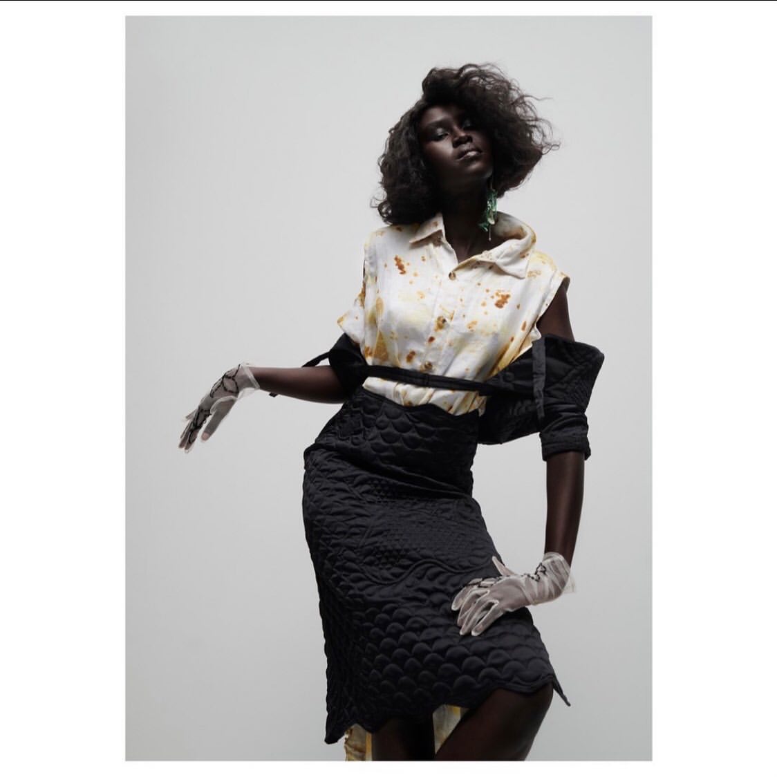 A dark-skinned woman poses against a pale background, wearing a patterned top, a black skirt, sheer gloves, and statement earrings in a studio shot.