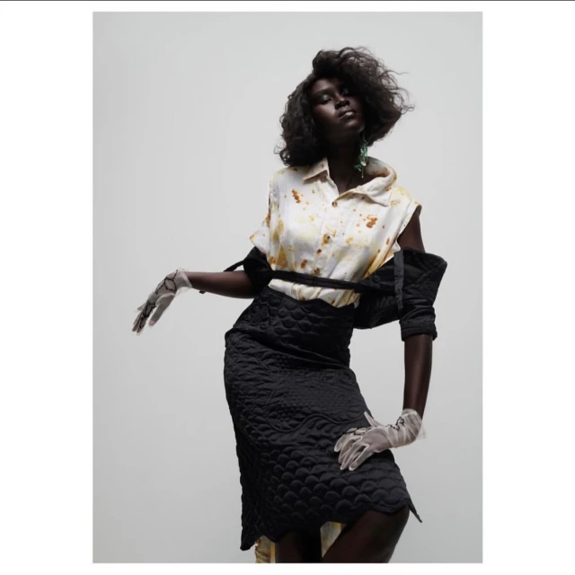 A dark-skinned woman poses against a pale background, wearing a patterned top, a black skirt, sheer gloves, and statement earrings in a studio shot.