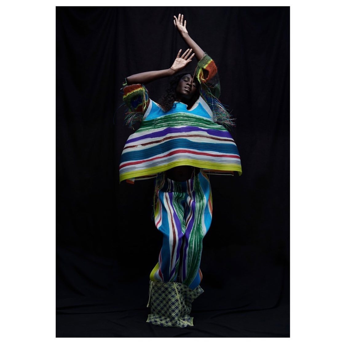 Black woman in colorful striped pleated top and trousers posing against a dark background