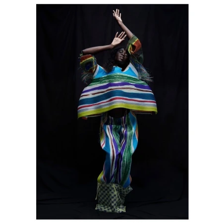 Black woman in colorful striped pleated top and trousers posing against a dark background