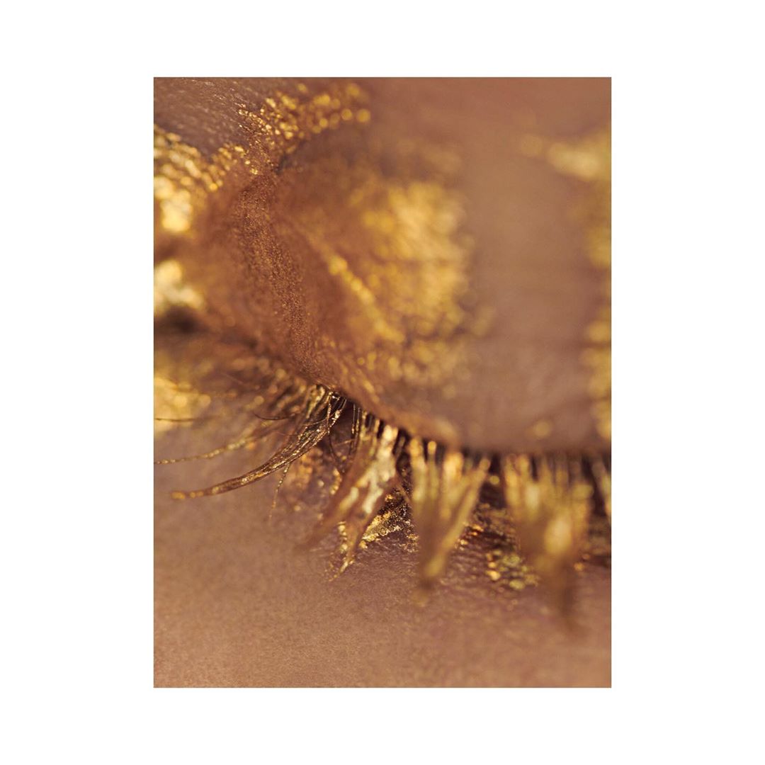 Close-up of a woman's eye adorned with shimmering gold pigment, catching the light with its textured, metallic finish and striking aesthetic.