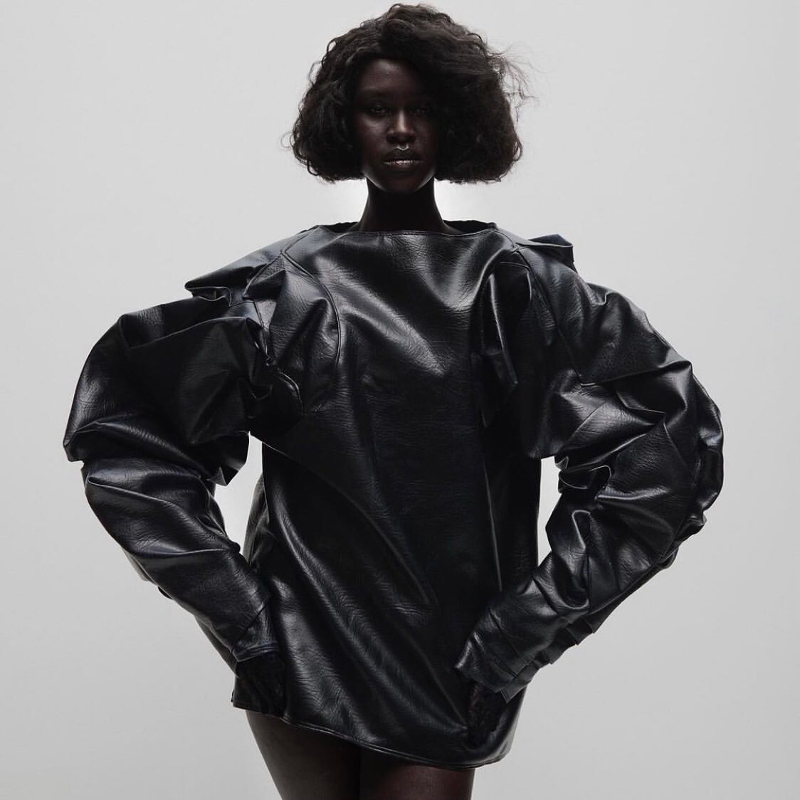 A dark-skinned woman poses in a studio wearing a black, oversized, long-sleeved top with statement puffy sleeves against a pale gradient backdrop.