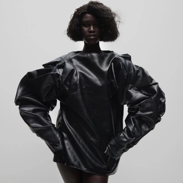 A dark-skinned woman poses in a studio wearing a black, oversized, long-sleeved top with statement puffy sleeves against a pale gradient backdrop.