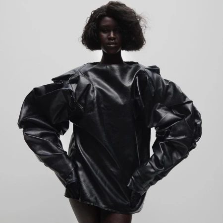 A dark-skinned woman poses in a studio wearing a black, oversized, long-sleeved top with statement puffy sleeves against a pale gradient backdrop.