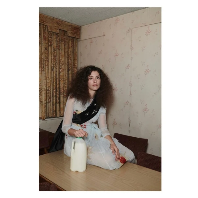 A young woman with dark curly hair sitting on a table indoors with a milk carton wearing a light-colored patterned dress and black sash.